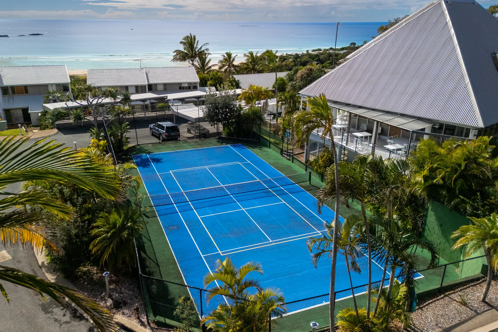 The blue tennis and pickle ball court at Pandanus Palms Resort, ringed by palms with the ocean beyond